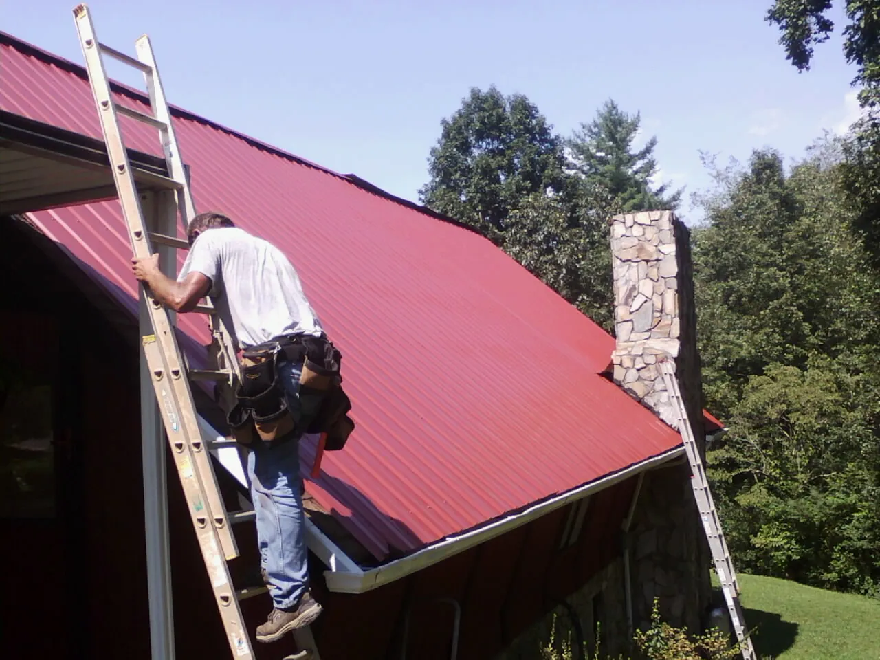 Roofer on a ladder beside a red metal roof and stone chimney — Soffit & Fascia Repair by Roofing Brothers in Wisconsin Dells